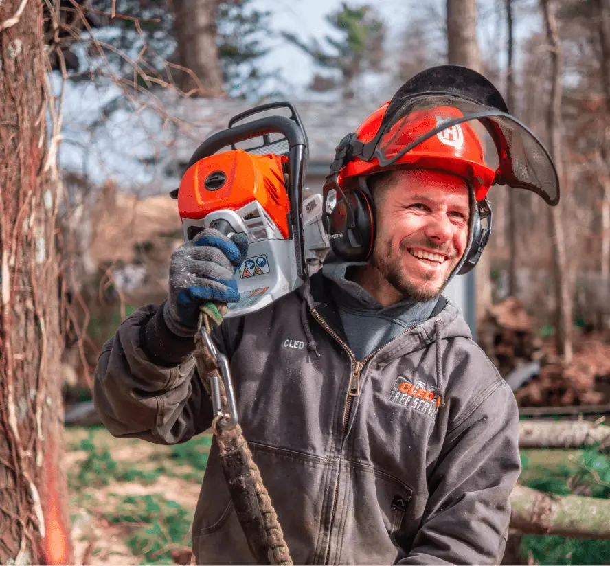 Cled Tree Service employee smiling while holding a chainsaw in a forested area, wearing protective gear and a helmet.