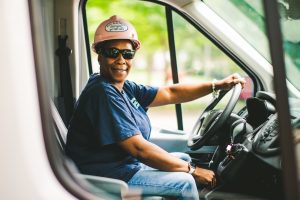 Marquita Kelley, wearing a pink hard hat and sunglasses, sits in the driver’s seat of a work van, smiling at the camera.